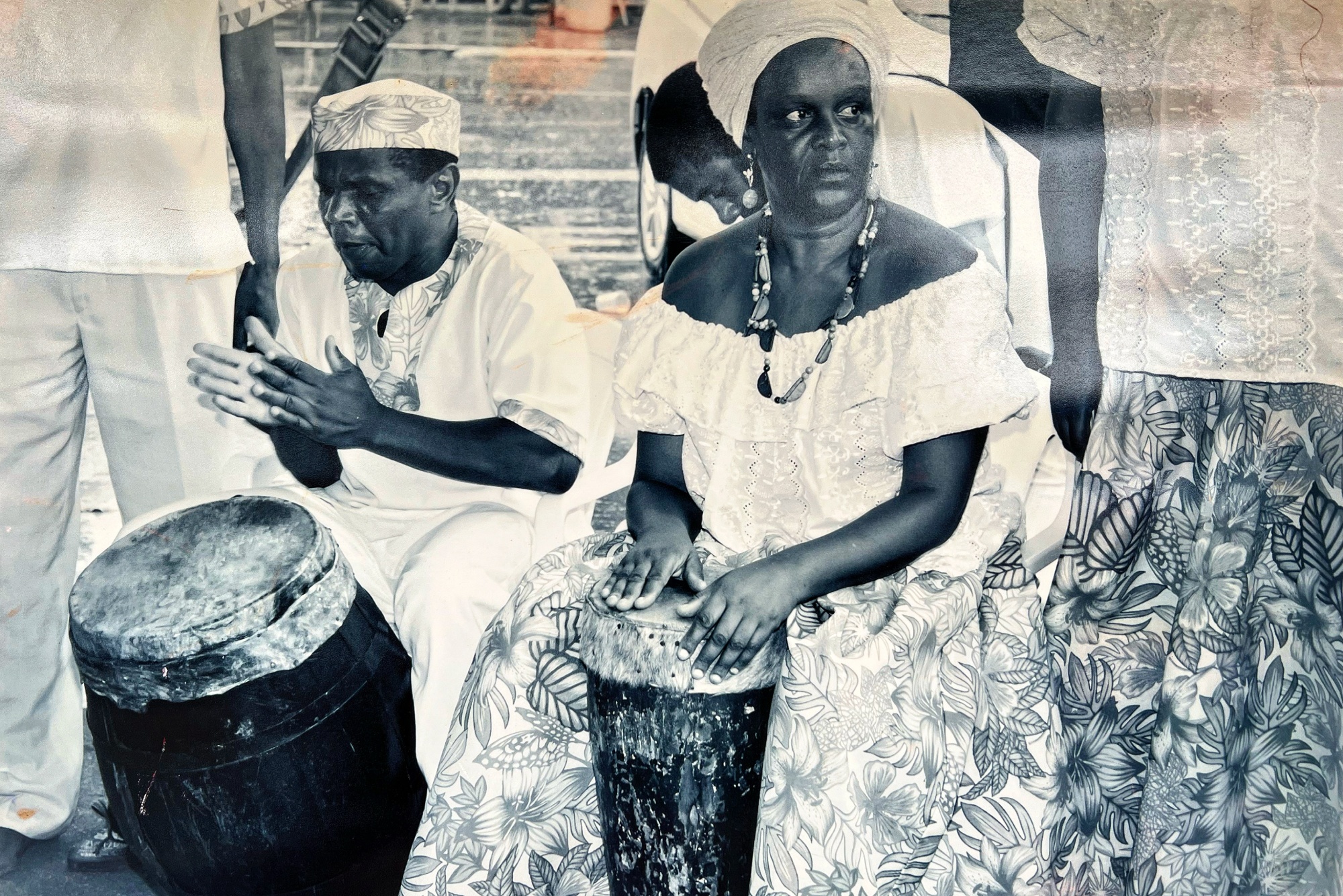 Mestra Meméia, Jongo de Pinheiral Foto 3 - Mestra Meméia, Jongo de Pinheiral • Acervo pessoal Descritivo: fotografia antiga, em preto e branco, com uma mulher negra sentada, de saia longa estampada florida e blusa branca, colar e turbante na cabeça, tocando tambor que está preso entre as suas pernas. Ao lado, um homem negro, de veste branca com detalhes no mesmo tecido estampado da saia e chapéu africano Kufi, também guarda um tambor entre as pernas e bate palmas.