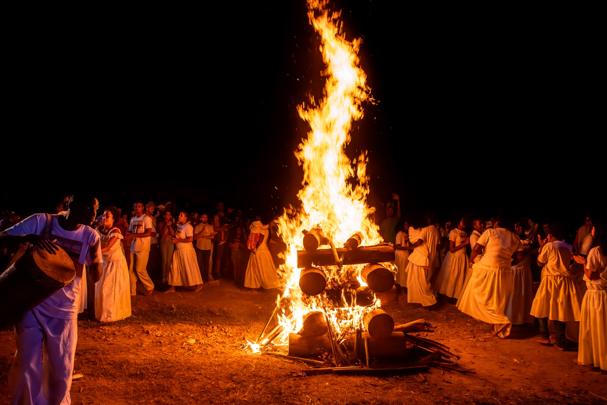 Roda de Jongo do Quilombo São José na Festa de Maio, em 2024, à noite em torno da fogueira. O chão é de barro e em torno da cena está escuro. Homens e mulheres se vestem todos de branco e estão de pés descalços. Um deles toca tambor enquanto os demais batem palmas.